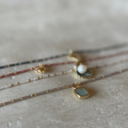 A close-up, shallow depth-of-field shot of several delicate beaded strands and pendants arranged on a textured light beige surface. The jewelry includes gold-toned chains with small gold beads, a pearl pendant, a dark fan-shaped charm, and a square pendant with a pale blue-gray stone in a gold bezel.
