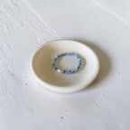 A beaded stretch bracelet with light blue and white beads, displayed inside a small, round, speckled ceramic dish on a textured white surface.