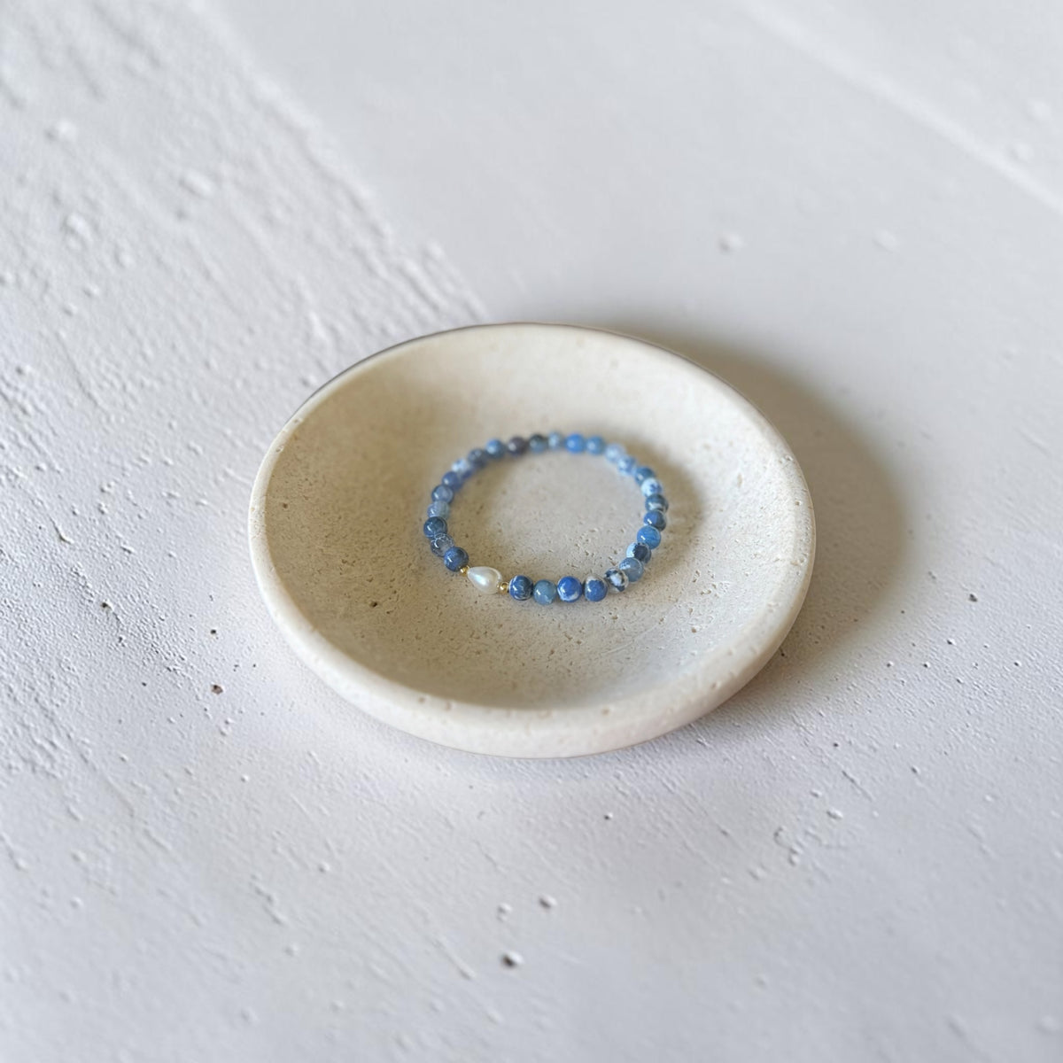 A beaded stretch bracelet with light blue and white beads, displayed inside a small, round, speckled ceramic dish on a textured white surface.