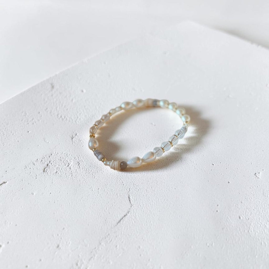 A delicate beaded stretch bracelet with small iridescent beads and a few pale gold spacer beads, resting on a textured white surface.