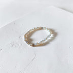 A delicate beaded stretch bracelet with small iridescent beads and a few pale gold spacer beads, resting on a textured white surface.