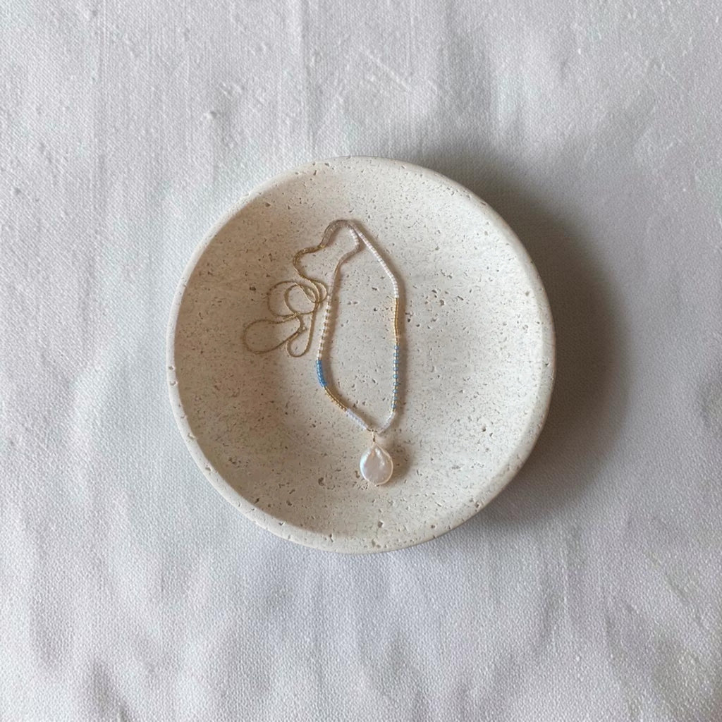 A delicate beaded necklace with a teardrop pendant rests inside a small, round, speckled ceramic dish on a textured white fabric background.