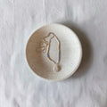 A delicate beaded necklace with a teardrop pendant rests inside a small, round, speckled ceramic dish on a textured white fabric background.