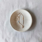 A delicate beaded necklace with a teardrop pendant rests inside a small, round, speckled ceramic dish on a textured white fabric background.