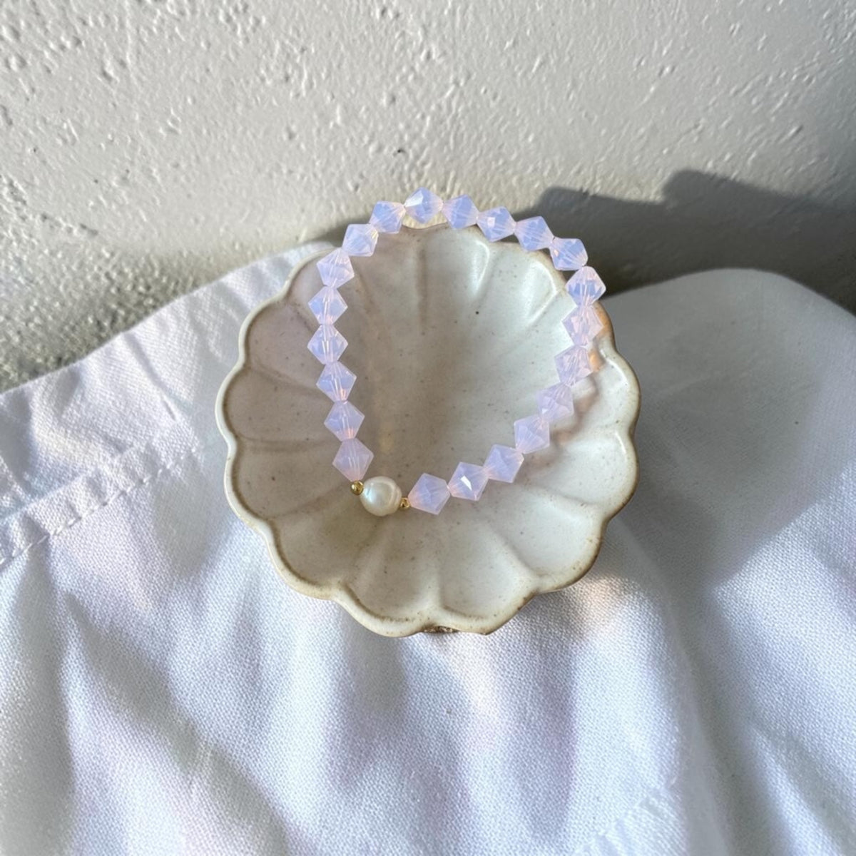 A beaded stretch bracelet with faceted pale lavender-purple beads and a single white pearl, displayed in a scalloped ceramic dish on white fabric.
