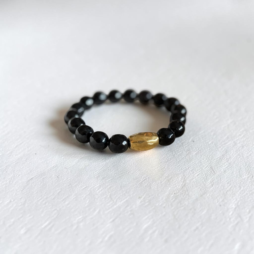A beaded stretch bracelet featuring glossy black round beads and a single textured, metallic gold oval accent bead, displayed on a textured white surface.