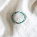 A thin turquoise blue beaded bracelet with a small iridescent bead accent, resting on a textured white fabric background.