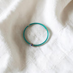 A thin turquoise blue beaded bracelet with a small iridescent bead accent, resting on a textured white fabric background.