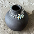 A beaded bracelet with clear faceted beads, a white pearl-like bead, and light blue stone beads with small gold accents, displayed on a small matte black vase against a textured concrete background.