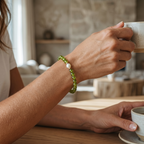 A person wearing a green beaded bracelet with a white pearl, holding a ceramic mug at a wooden table in a softly blurred indoor setting.