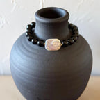 A beaded bracelet with glossy black round beads and a central iridescent white shell-like charm, displayed around a matte black ceramic vase against a light textured wall.