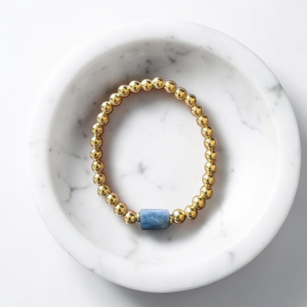 A gold beaded stretch bracelet with a single blue rectangular stone accent, displayed inside a white marble bowl on a light gray surface.