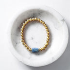 A gold beaded stretch bracelet with a single blue rectangular stone accent, displayed inside a white marble bowl on a light gray surface.