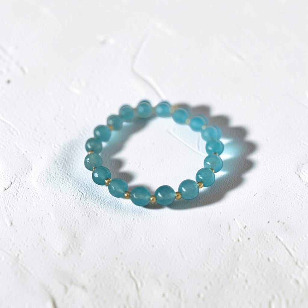 A beaded stretch bracelet made of translucent light blue-green round beads separated by small gold spacer beads, photographed on a textured white surface.