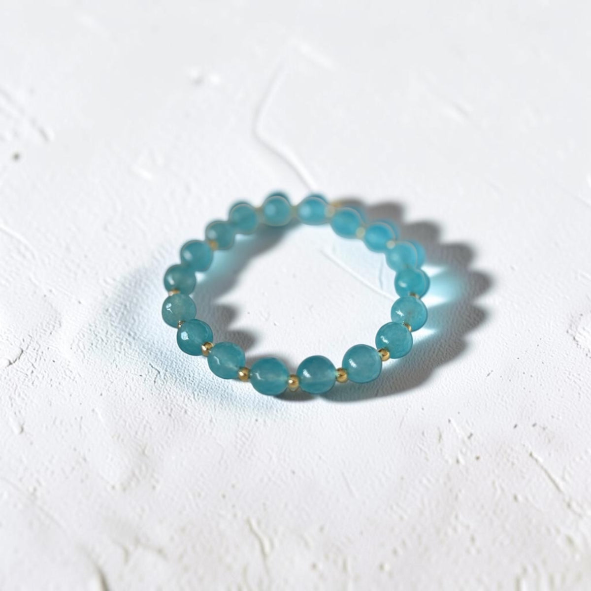 A beaded stretch bracelet made of translucent light blue-green round beads separated by small gold spacer beads, photographed on a textured white surface.