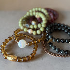 A group of beaded bracelets arranged on a neutral surface. The set includes translucent amber-yellow beads with a white iridescent bead, light olive-green beads, dark charcoal gray beads, and a rose-gold chain bracelet.