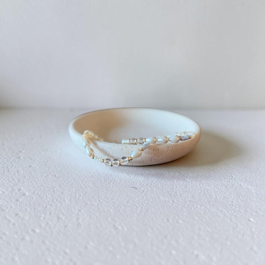 A delicate beaded accessory with small white pearls and clear beads draped over a small beige ceramic dish on a textured white surface.