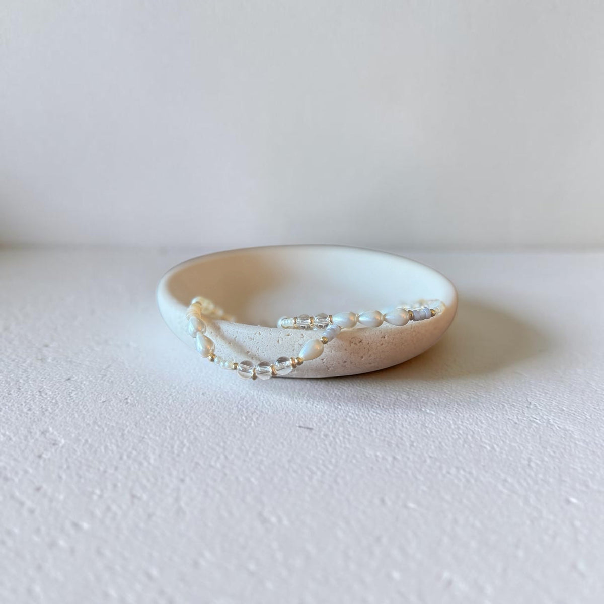 A delicate beaded accessory with small white pearls and clear beads draped over a small beige ceramic dish on a textured white surface.