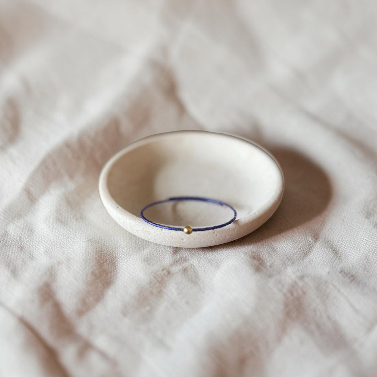 A delicate thin purple blue wire bracelet with a small gold bead, displayed inside a small white ceramic dish on a textured beige linen fabric background.
