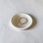 A delicate peach-beige beaded bracelet with a small white pearl accent, displayed on a small speckled ceramic dish against a textured light gray background.