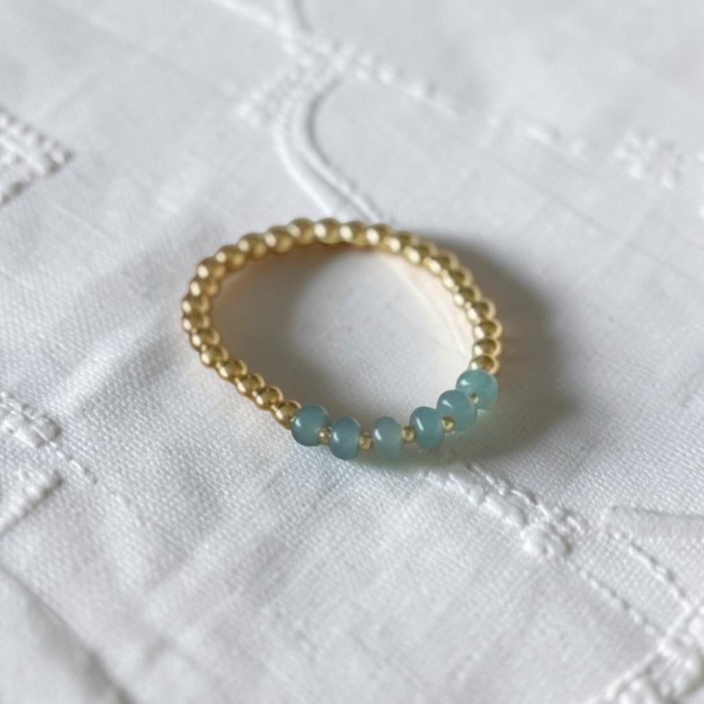 A gold beaded bracelet with five light blue round beads, resting on a textured white fabric background.