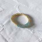 A gold beaded bracelet with five light blue round beads, resting on a textured white fabric background.