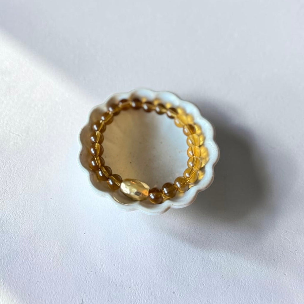 A beaded braceletwith translucent amber-brown beads and a central faceted clear stone, displayed on a small white scalloped dish against a light gray background.