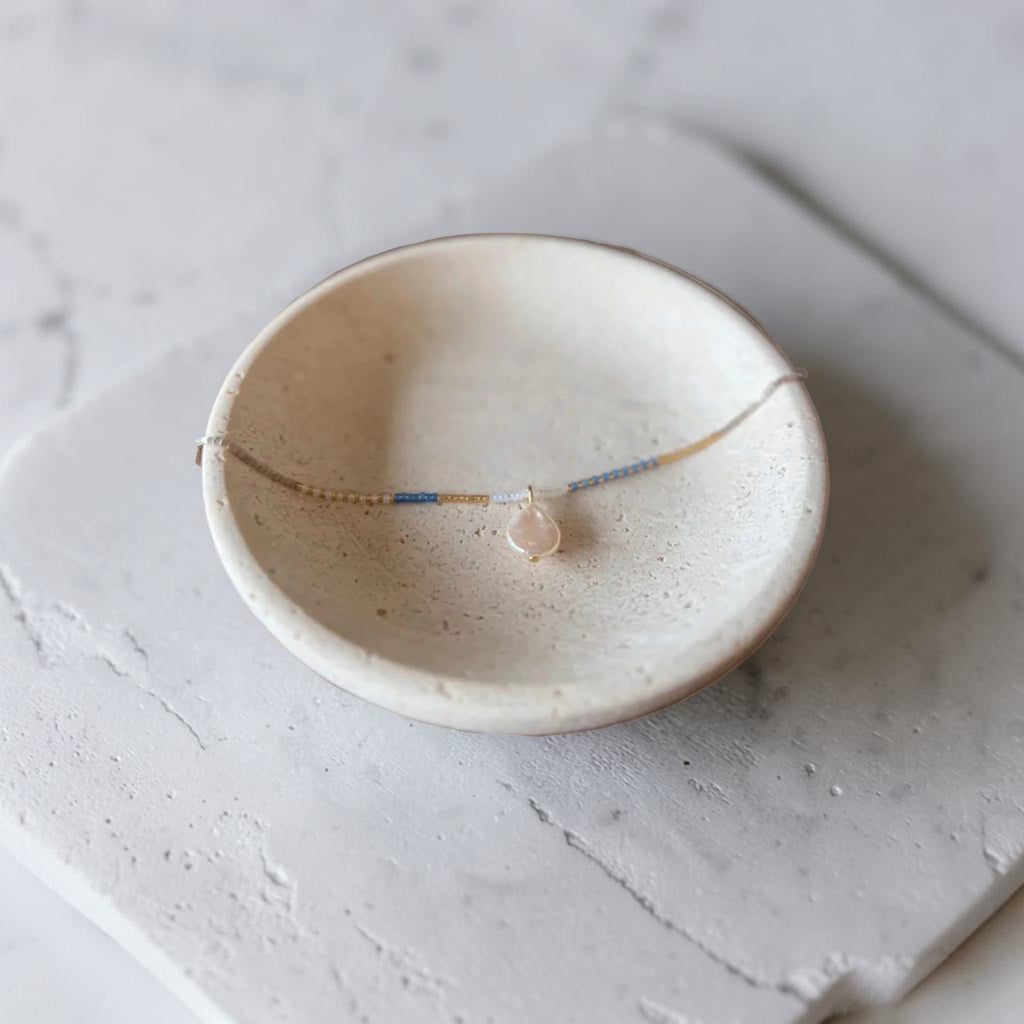 A delicate beaded necklace with a small peach-colored bead pendant, displayed inside a speckled cream ceramic dish on a light gray stone surface.