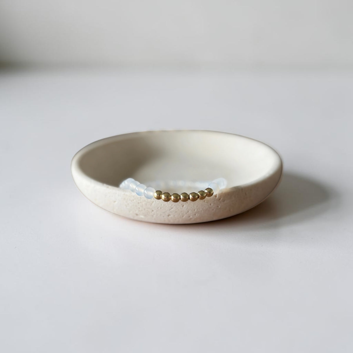 A small round ceramic dish in a warm off-white or cream glaze, holding a delicate beaded bracelet with gold-toned beads and a pale iridescent bead, set against a minimalist light gray background.