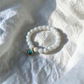 A beaded stretch bracelet made of white beads with a small gold-toned charm featuring a round turquoise-blue stone, resting on a textured white fabric surface.