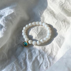 A beaded stretch bracelet made of white beads with a small gold-toned charm featuring a round turquoise-blue stone, resting on a textured white fabric surface.