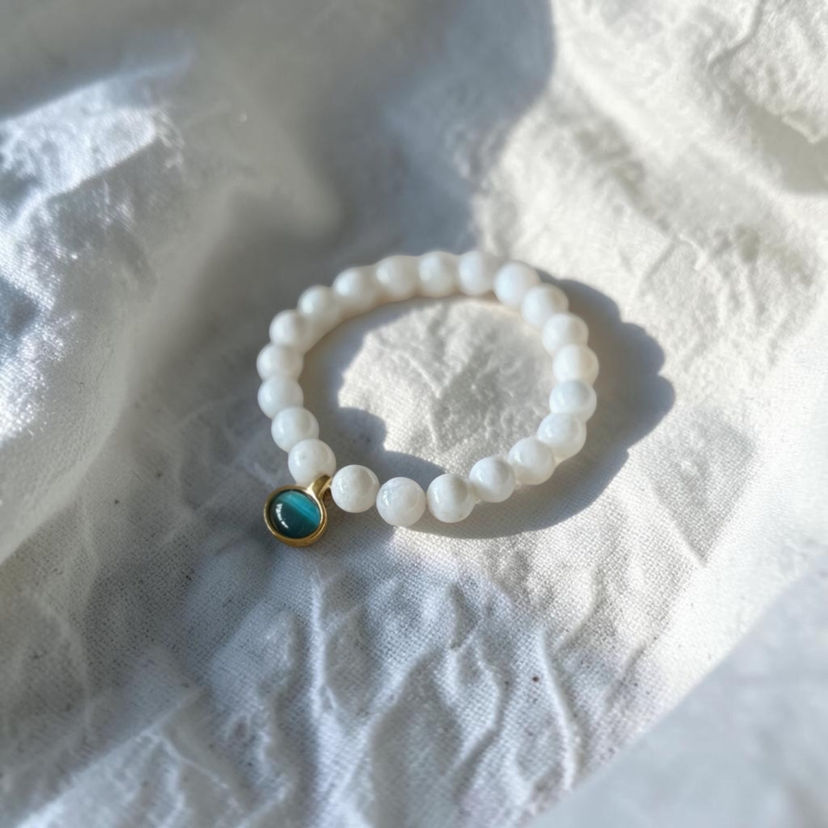 A beaded stretch bracelet made of white beads with a small gold-toned charm featuring a round turquoise-blue stone, resting on a textured white fabric surface.