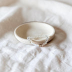 A delicate  clear stone bracelet rests inside a small, round, speckled cream ceramic dish on a textured white linen fabric background.
