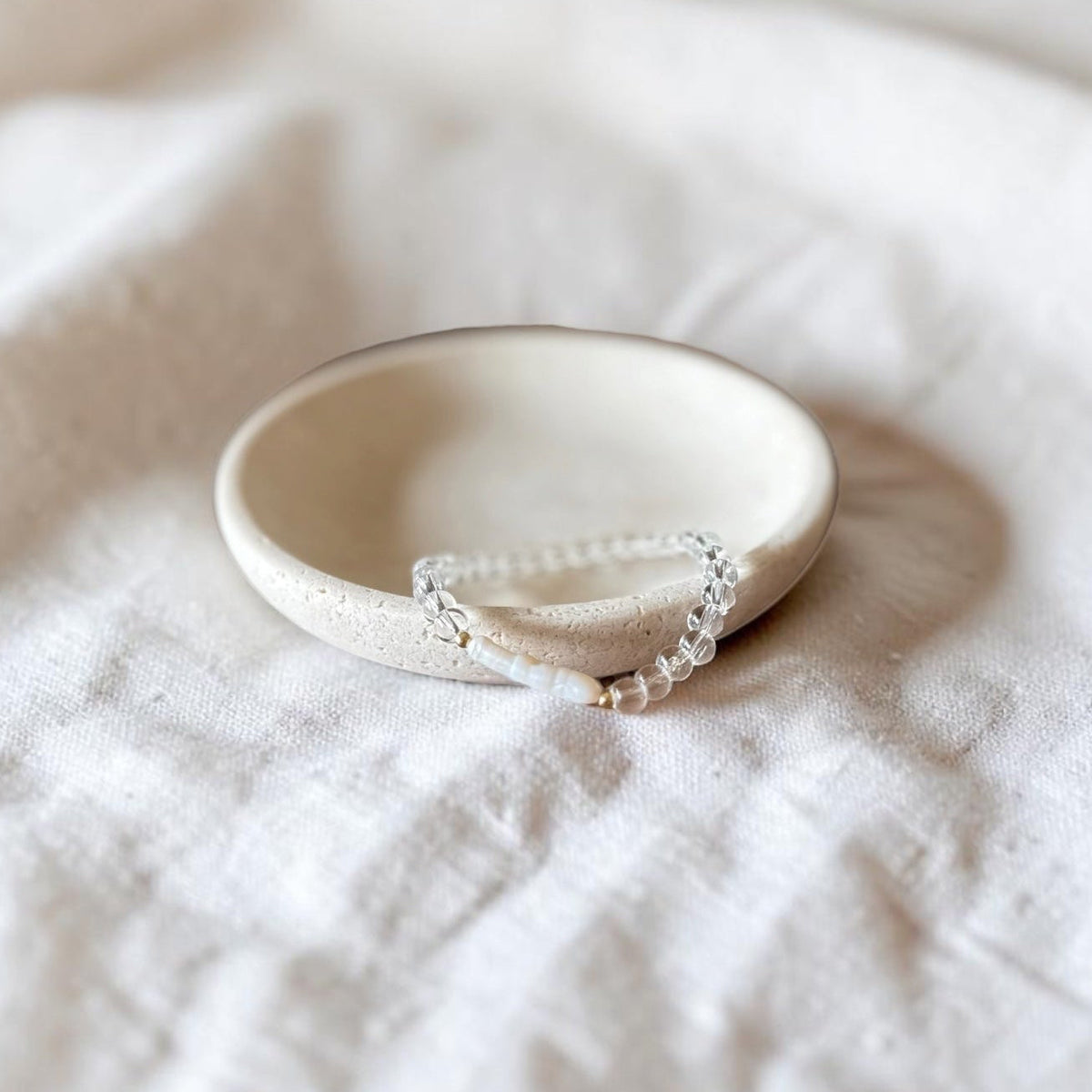 A delicate  clear stone bracelet rests inside a small, round, speckled cream ceramic dish on a textured white linen fabric background.
