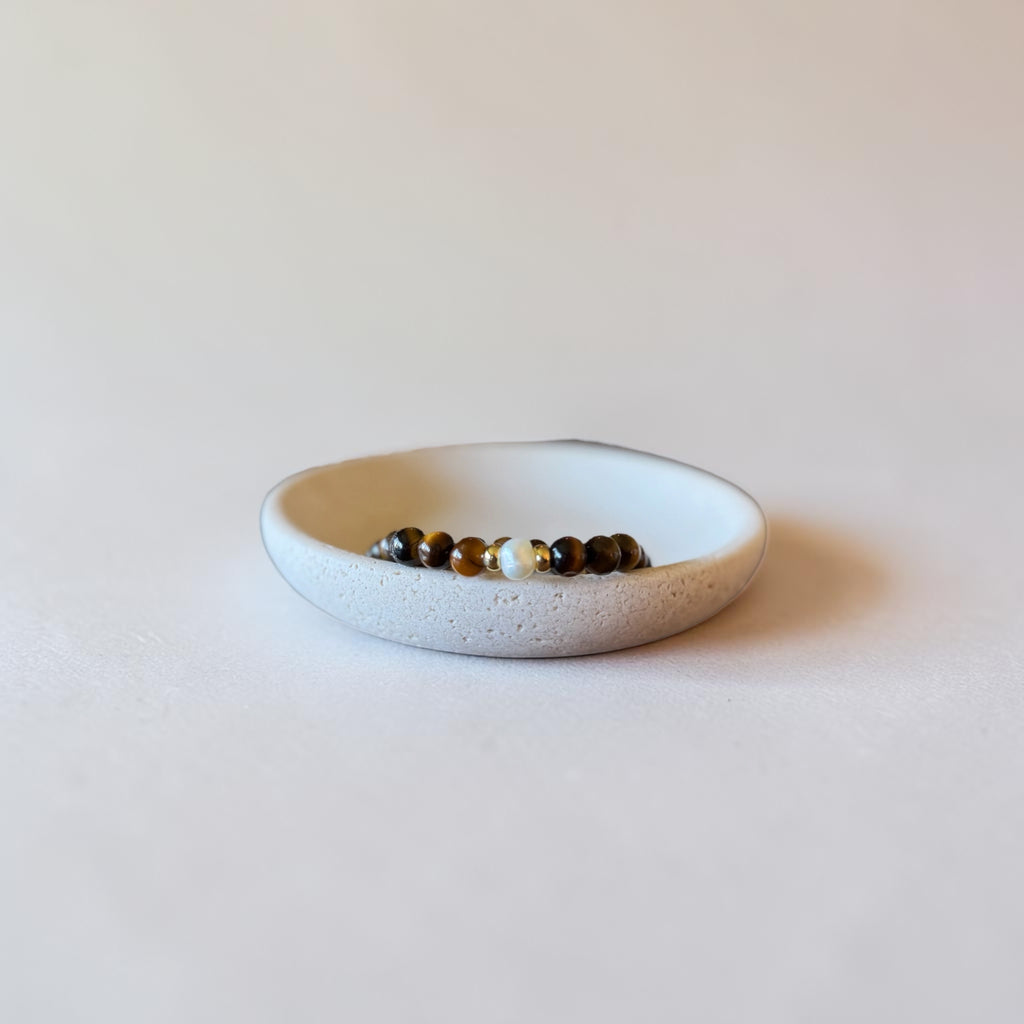 A small beaded stretch bracelet with brown, black, and white beads resting inside a shallow, speckled off-white ceramic dish on a light beige background.