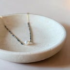 A delicate necklace with a single white pearl resting on a textured, shallow ceramic dish.