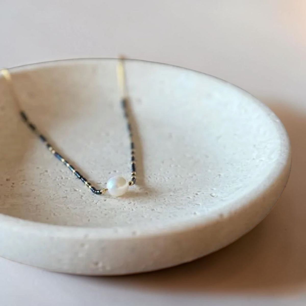 A delicate necklace with a single white pearl resting on a textured, shallow ceramic dish.