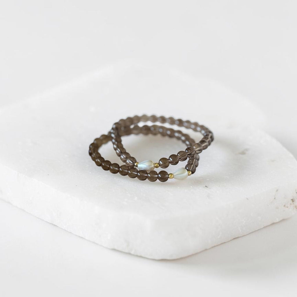 Two beaded wrap rings in a dark brown color, featuring a small iridescent white bead accent, displayed on a white stone block against a white background.