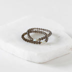 Two beaded wrap rings in a dark brown color, featuring a small iridescent white bead accent, displayed on a white stone block against a white background.