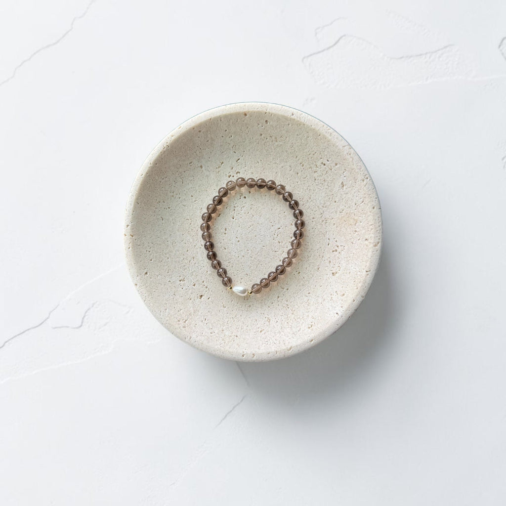 A beaded bracelet with small brown beads and a silver clasp, displayed inside a round, speckled ceramic dish on a light gray textured surface.