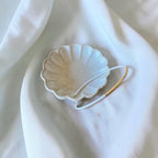 A small scalloped ceramic dish in a warm off-white glaze with subtle speckles, holding a thin white wire bracelet. It is sitting on the dish to show how the bracelet opens, styled on draped white fabric.