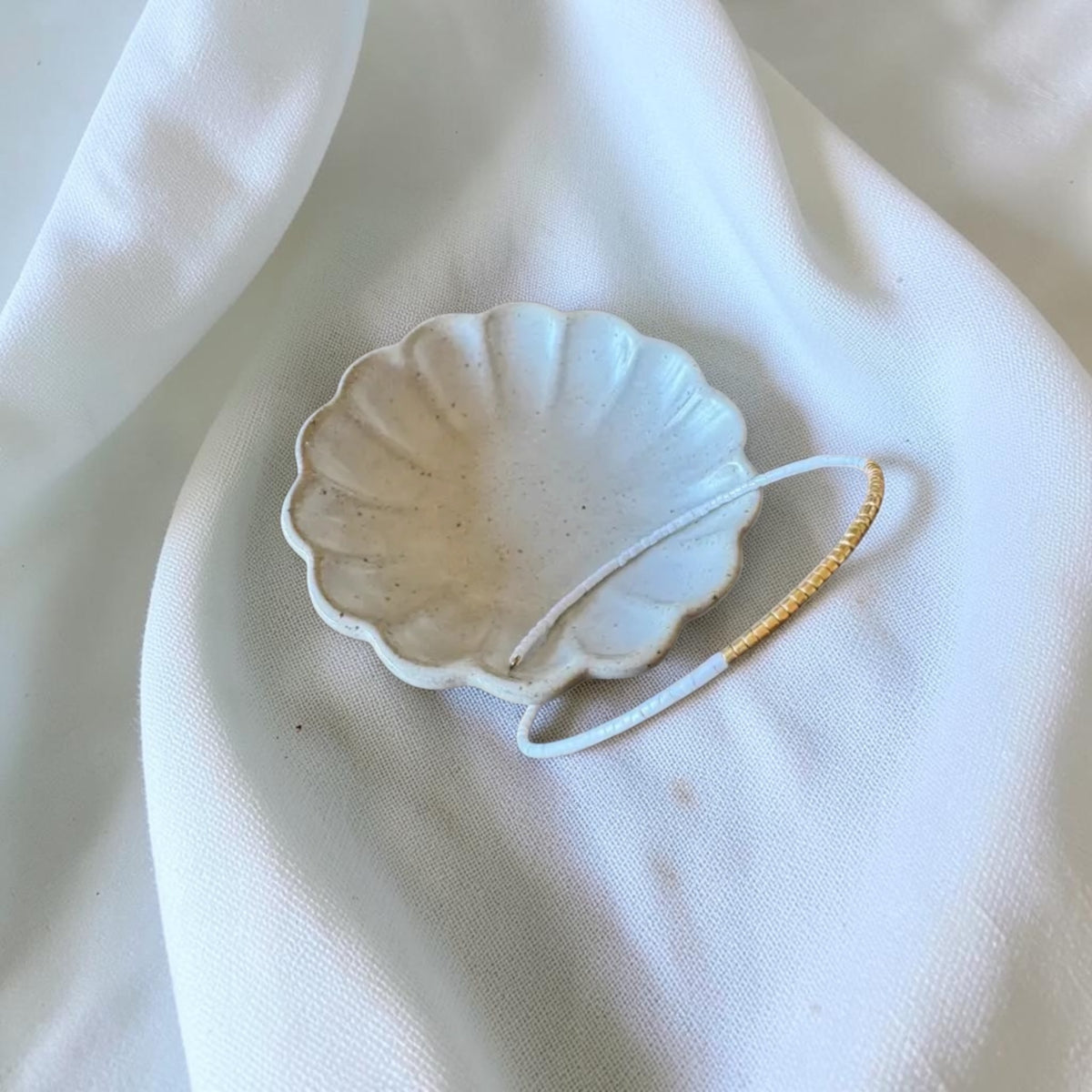 A small scalloped ceramic dish in a warm off-white glaze with subtle speckles, holding a thin white wire bracelet. It is sitting on the dish to show how the bracelet opens, styled on draped white fabric.