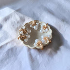 A small scalloped cream dish holding six pearl earrings with gold-toned centers, photographed on a soft white fabric surface.