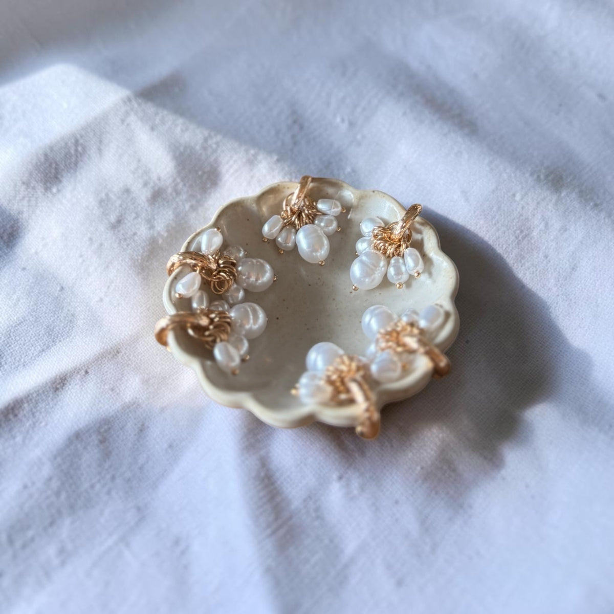 A small scalloped cream dish holding six pearl earrings with gold-toned centers, photographed on a soft white fabric surface.