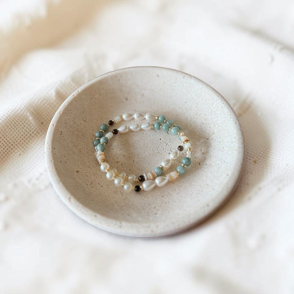 A beaded stretch bracelet with white pearls, light blue beads, and small black beads, displayed on a small speckled ceramic dish over a soft white fabric background.