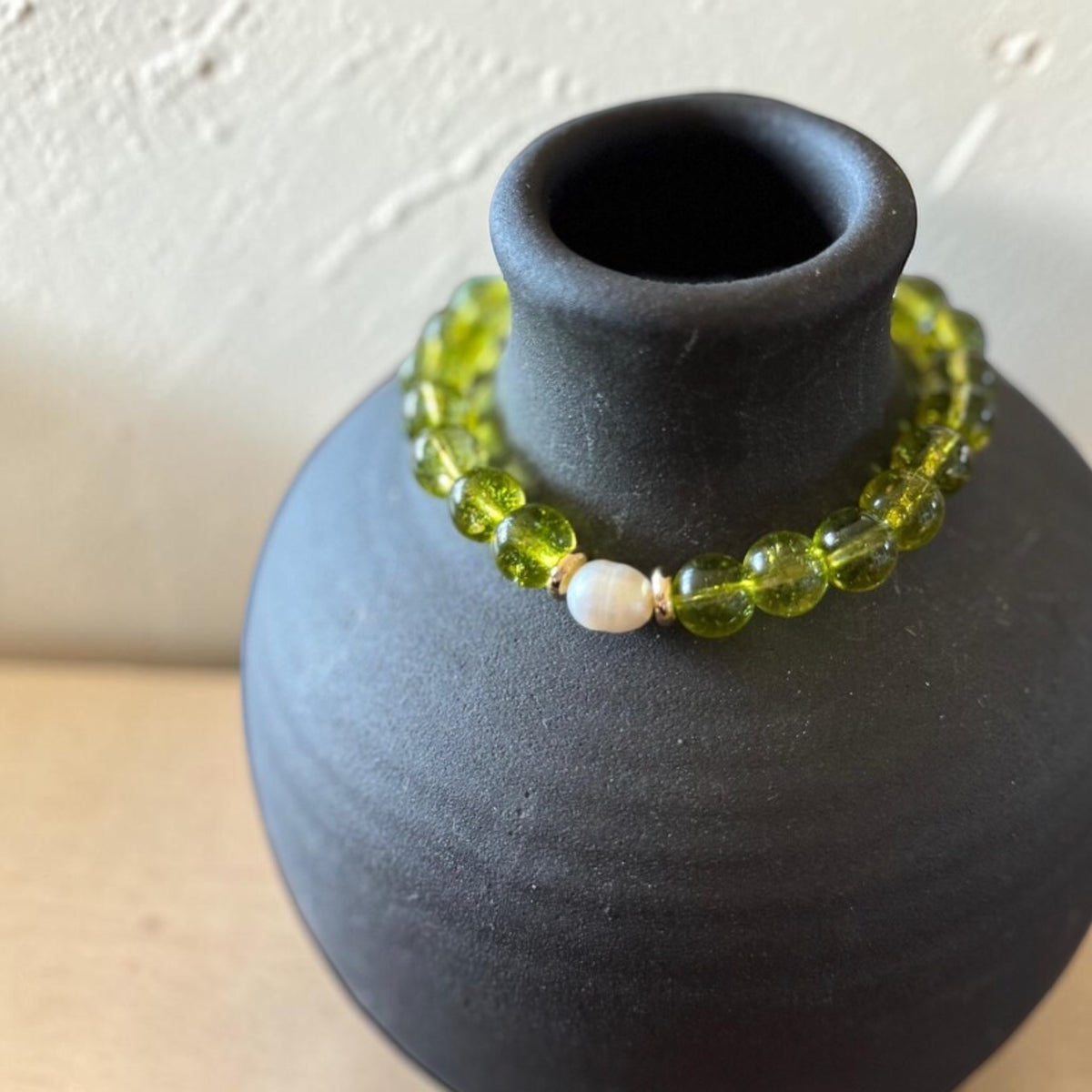 A beaded bracelet with translucent olive green round beads and a single white pearl-like bead, displayed on a matte black ceramic vase against a light textured wall.