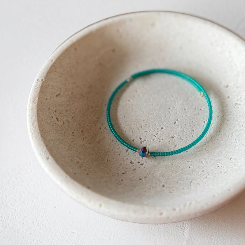 A thin turquoise green beaded bracelet with a small iridescent bead, displayed inside a textured beige ceramic bowl on a white surface.