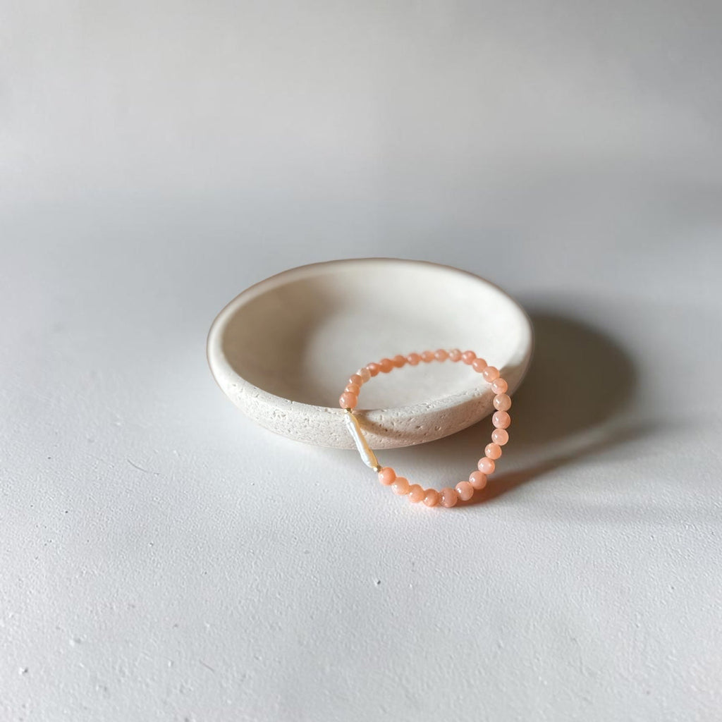 A peach-colored beaded bracelet with a white cord loop, resting on a small, textured off-white ceramic dish against a neutral light gray background.