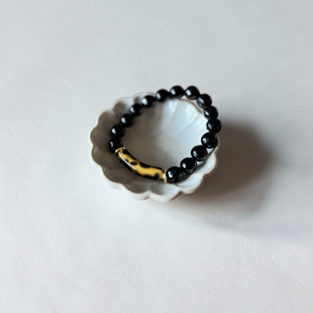 A beaded stretch bracelet with glossy black round beads and a polished gold-toned accent piece, displayed on a small white scalloped dish against a light gray background.
