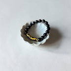 A beaded stretch bracelet with glossy black round beads and a polished gold-toned accent piece, displayed on a small white scalloped dish against a light gray background.
